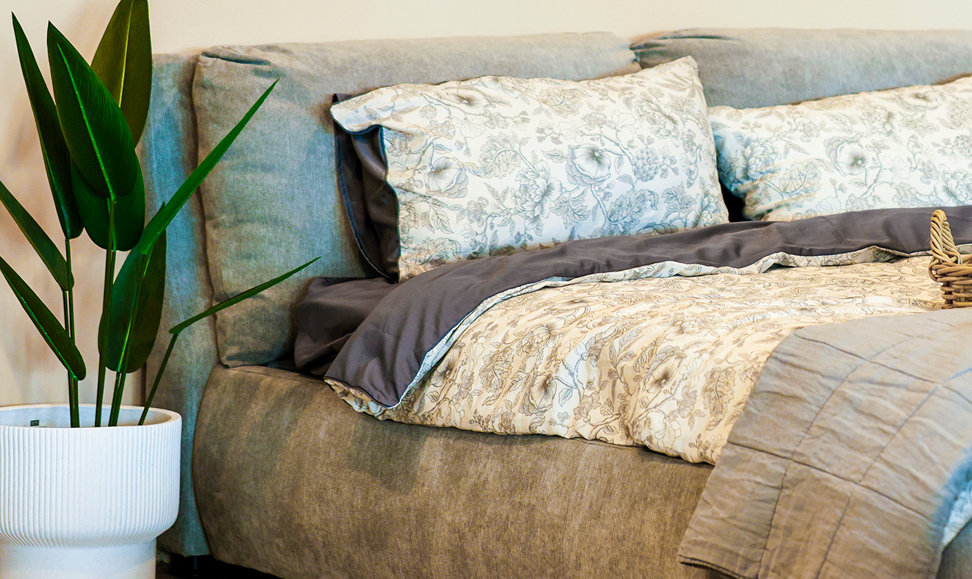 Upholstered bed with neutral fabric frame, styled with patterned bedding and cushions beside a potted plant.